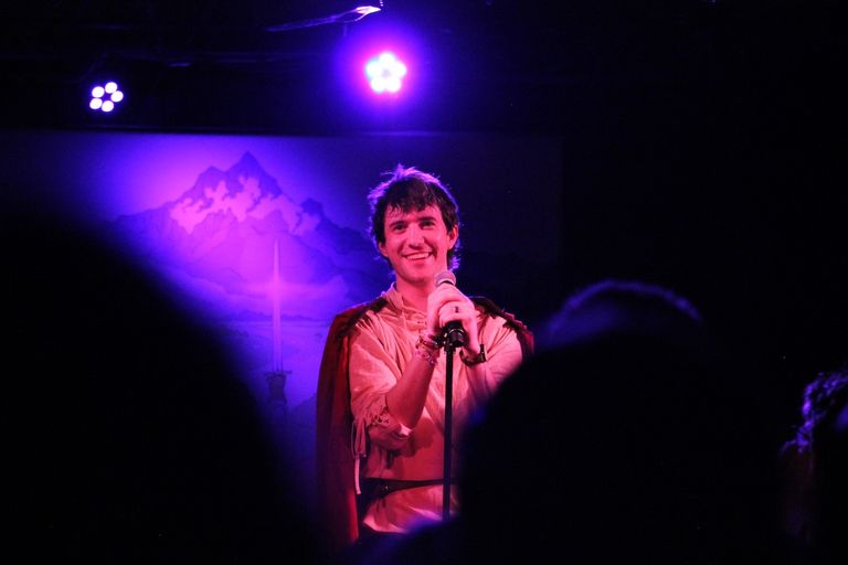 Musician Naethan Apollo smiles into a microphone on stage, wearing a tunic and cape under purple lighting during a live concert performance.